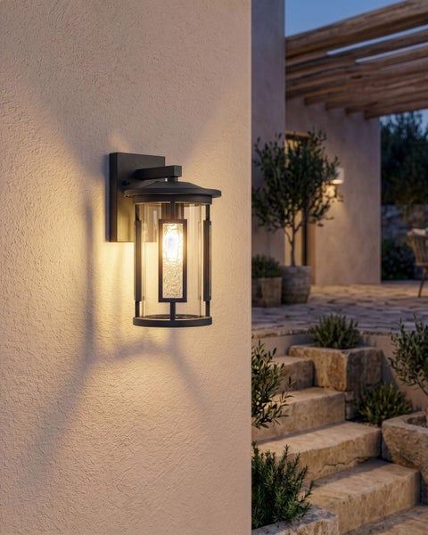 Moderne Außenwandleuchte in Laternenform an einer strukturierten Fassade, warmes Licht auf einer mediterranen Terrasse mit Steintreppe und Pflanzen.