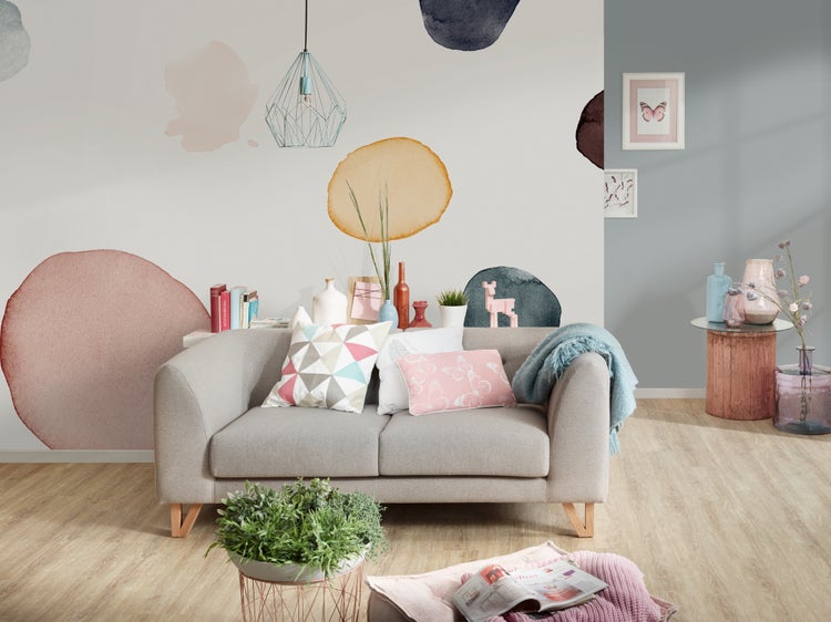 Modernes Wohnzimmer mit grauem Zweisitzer-Sofa, Wandgestaltung mit abstrakten Aquarell-Kreisen, geometrischer Pendelleuchte und Dekokissen.