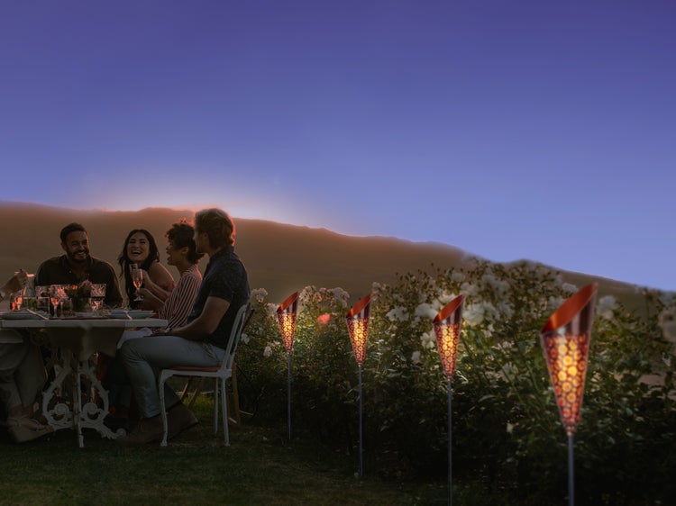 Szene im Freien mit einer Gruppe von Menschen, die an einem Tisch sitzen und Gartenfackeln, die für eine gemütliche Atmosphäre sorgen.