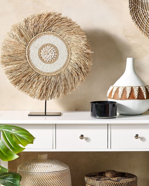 Dekoratives Stillleben auf einem weissen Tisch mit einer Vase, einer schwarzen Schale, Körben und einer Wanddekoration aus Rattan.
