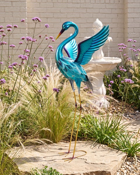 Blaue Gartenfigur Reiher aus Metall mit ausgebreiteten Flügeln in einem hellen Garten mit Ziergräsern, Verbena und einem Springbrunnen aus Stein.
