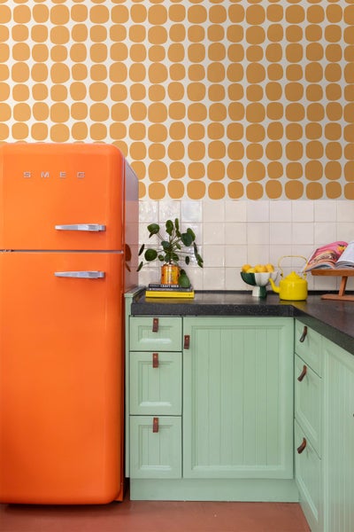 Hellgrüne Küchenschränke mit Ledergriffen, dunkle Arbeitsplatte, weißer Fliesenspiegel, orange gemusterte Tapete und Smeg Logo auf Kühlschrank.