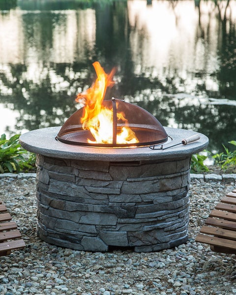 Runde Feuerstelle in Steinoptik mit Funkenschutzhaube und brennendem Feuer auf einer Kiesfläche am Wasser.