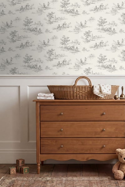 Vliestapete im Toile-de-Jouy-Stil mit ländlichen Szenen über einer weißen Wandvertäfelung und einer Holzkommode in einem hellen Kinderzimmer.