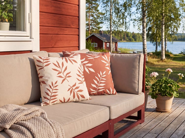 Gartensofa mit Kissen mit Blattmuster auf einer Holzterrasse mit Blick auf einen See und ein rotes Haus im Hintergrund.
