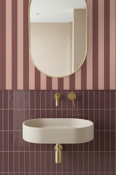 Modernes Badezimmer mit ovalem Waschbecken, goldfarbener Wandarmatur und ovalem Spiegel vor vertikalen Fliesen und gestreifter Tapete.