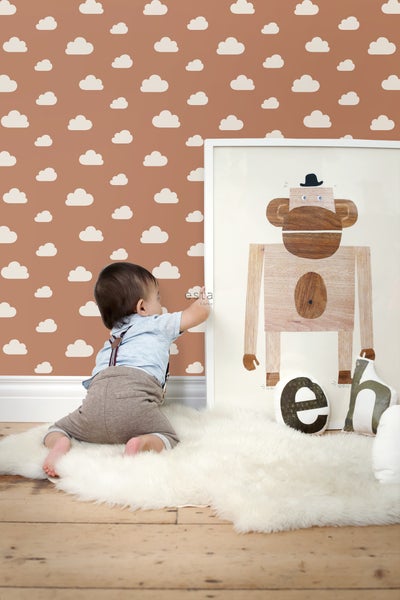 Baby vor einer Wand mit Wolkenmuster, einem gerahmten Affenbild und Kissen auf einem Teppich.