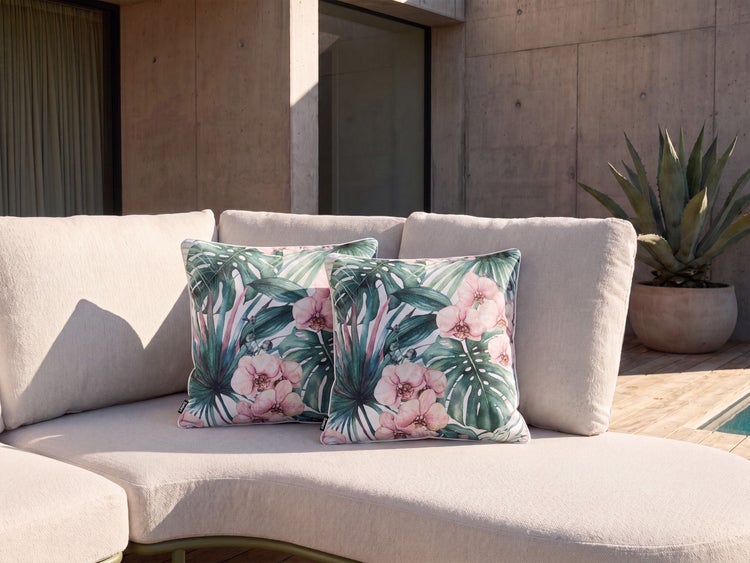 Zwei quadratische Zierkissen mit Blumen- und Blättermuster auf einem hellen Sofa im Freien vor einer Betonwand.