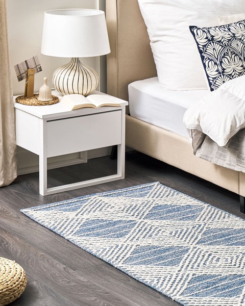 Hell eingerichtetes Schlafzimmer mit blau-weißem geometrischem Teppich auf dunklem Holzboden, beigem Polsterbett und weißem Nachttisch.