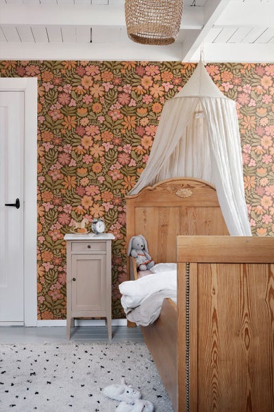 Kinderzimmer mit Retro-Blumentapete in Pink und Orange. Holzbett mit weißem Betthimmel, beiger Nachttisch und gewebte Deckenlampe.