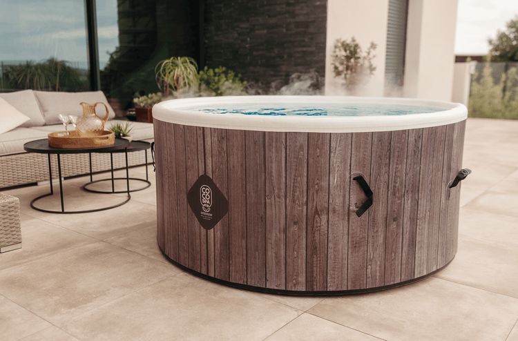 Runder aufblasbarer Whirlpool in Holzoptik mit CoCo Spa Logo auf einer hellen Terrasse mit Lounge-Möbeln und dampfendem Wasser.