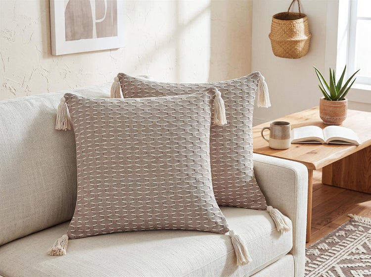 Zwei strukturierte Dekokissen mit Quasten auf einem Sofa in einem gemütlich eingerichteten Wohnzimmer.