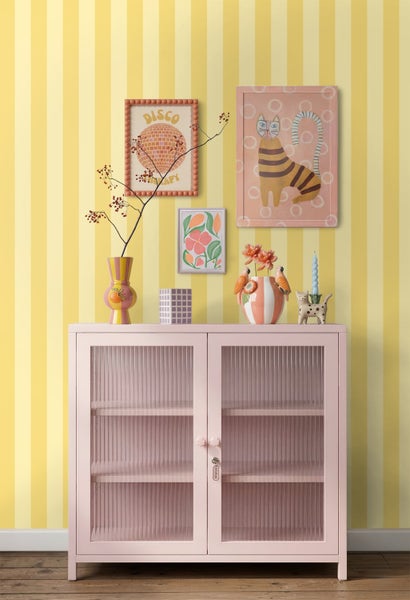 Vliestapete mit gelben und weißen Streifen hinter einer rosa Vitrine mit geriffeltem Glas und bunten Dekorationsartikeln.