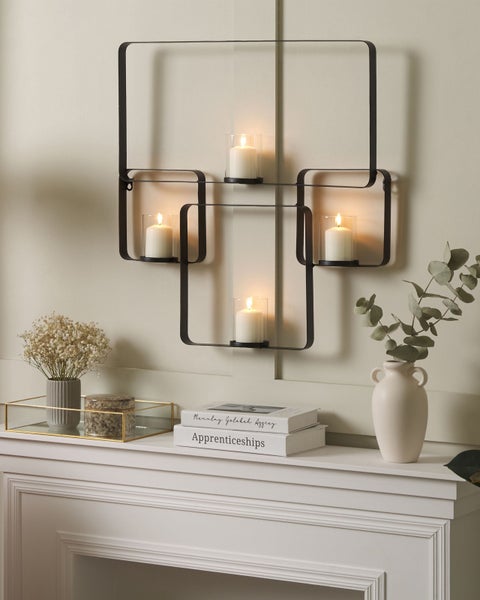 Wanddekoration mit Kerzenhalter aus Metall und Glaseinsätzen über einem Kaminsims mit Büchern und Dekorationen.