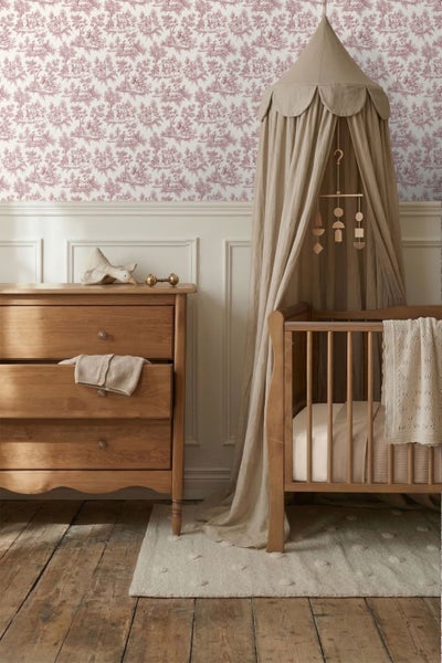 Kinderzimmer mit Toile-de-Jouy-Tapete, weißer Wandtäfelung, Holzkommode und Babybett mit Stoffhimmel auf Holzdielenboden.