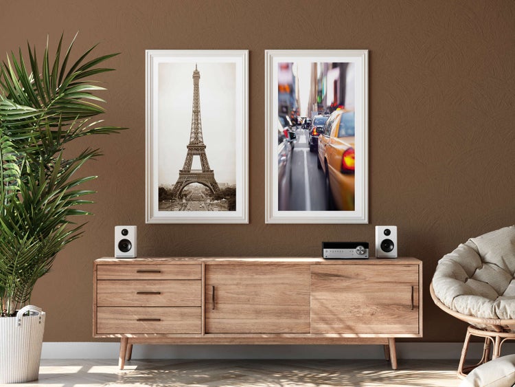 Helles Wohnzimmer mit zwei eingerahmten Bildern vom Eiffelturm und New York über einem Sideboard aus Holz mit Lautsprechern und einer Pflanze.