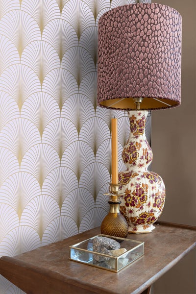 Szene mit einer Tischlampe mit gemustertem Lampenschirm, einer Kerze, Dekorationsgegenständen und einer Tapete mit geometrischem Muster.