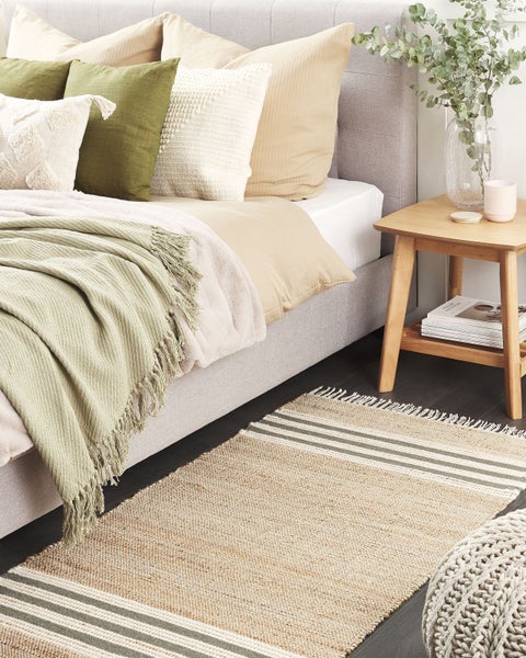 Modernes Schlafzimmer mit grauem Polsterbett, hellgrünem Überwurf, Dekokissen, Nachttisch aus Holz und einem gewebten Juteteppich.