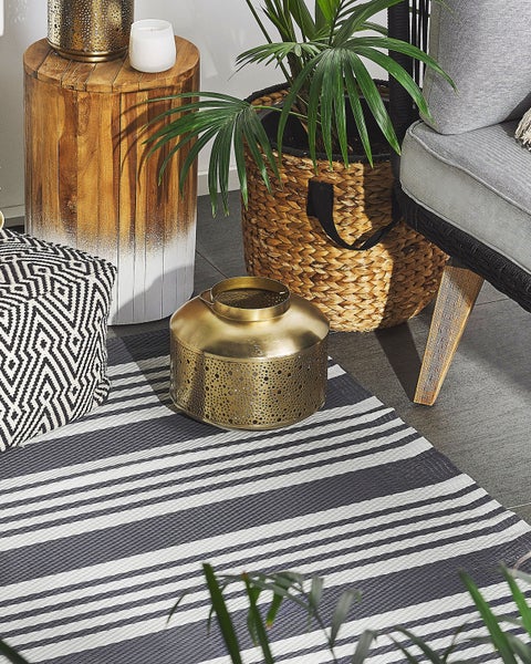 Wohnzimmerszene mit gestreiftem Teppich, goldener Metalllaterne, geometrischem Pouf, Holztisch und Pflanze im Flechtkorb.