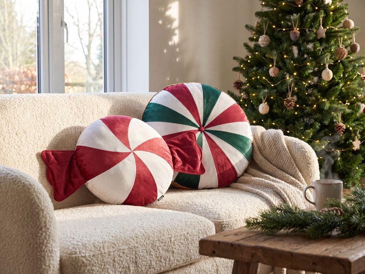 Zwei runde Weihnachts-Dekokissen im Bonbon- und Pfefferminz-Design auf einem hellen Sofa neben einem geschmückten Tannenbaum.