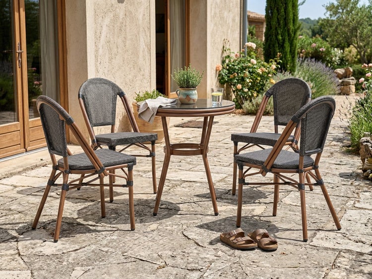 Gartenmöbel-Set bestehend aus einem runden Tisch mit Glasplatte und vier Bistrostühlen mit Geflecht auf einer Steinterrasse.