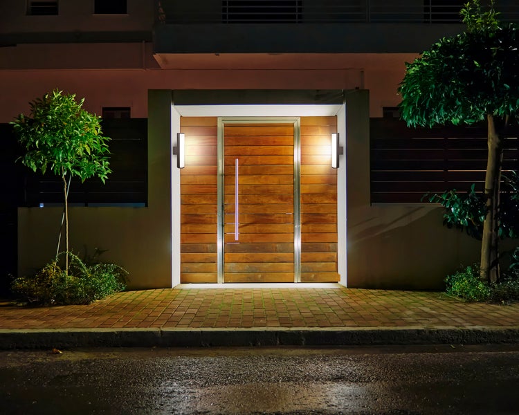 Außenansicht eines Hauses bei Nacht mit einer modernen Holztür, flankiert von zwei Außenwandleuchten und Bäumen