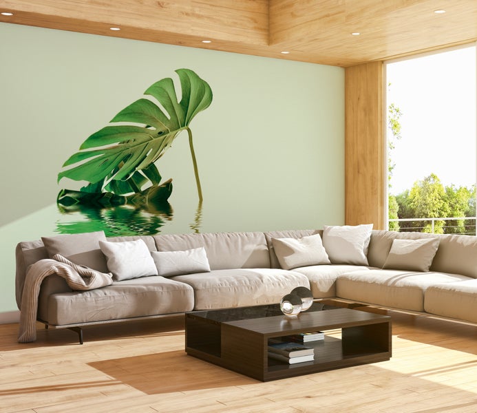 Helles Wohnzimmer mit einer Fototapete eines Monstera-Blattes im Wasser, großem Ecksofa, dunklem Couchtisch und Holzboden.