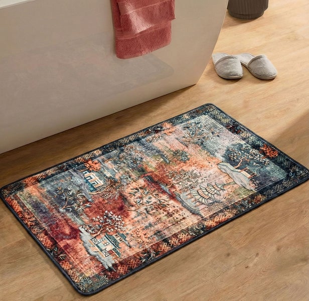 Rechteckige Badematte mit kunstvollem Landschaftsmuster auf Holzboden vor einer Badewanne mit Handtuch und Hausschuhen.