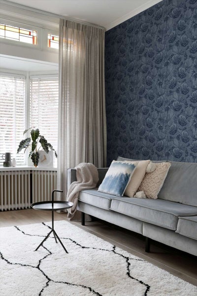 Wohnzimmer mit dunkler Blumentapete, Sofa mit Dekokissen, Teppich mit Muster und Beistelltisch vor einem Fenster mit Vorhängen.
