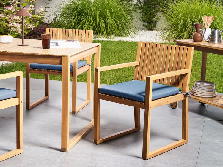 Modernes Gartenmöbel-Set aus hellem Holz mit Esstisch und Armlehnstühlen mit blauen Sitzkissen auf einer gefliesten Terrasse im Garten.