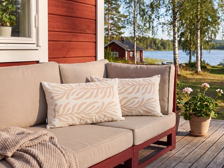 Gartensofa mit Zierkissen auf einer Holzterrasse vor einem Gartenhaus mit Blick auf einen See.