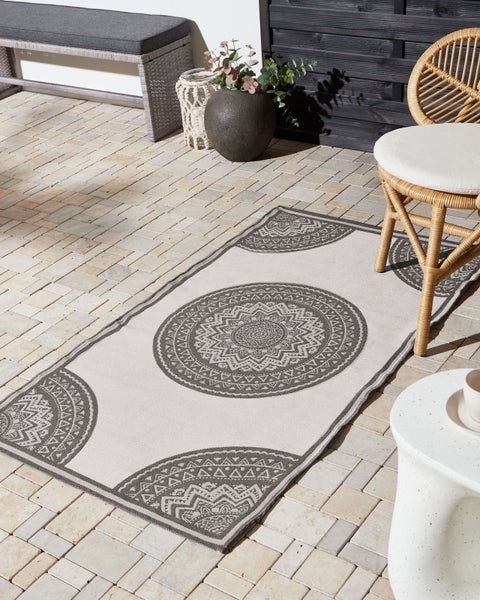 Heller Terrassenbereich mit Steinpflaster, einem rechteckigen Outdoor-Teppich mit Mandala-Mustern, Flechtmöbeln und einer Ziervase.