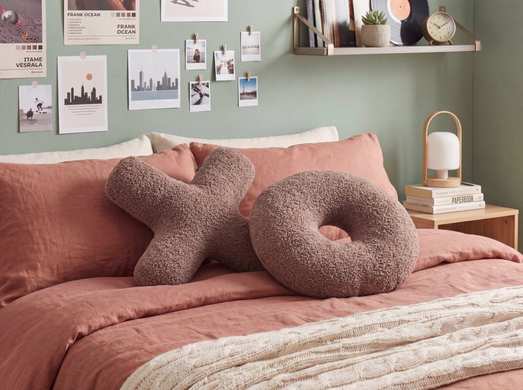 Hell gestaltetes Schlafzimmer mit Dekokissen in X- und O-Form aus strukturiertem Stoff auf einem Bett mit Leinenbettwäsche und Strickdecke.