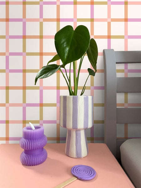 Modernes Raumdesign mit bunter karierter Tapete, einer Monstera-Pflanze in einer gestreiften Keramikvase und einer lila Kerze auf einem rosa Tisch.