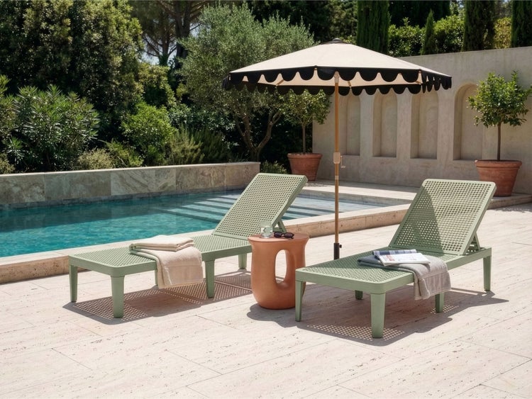 Zwei grüne perforierte Sonnenliegen mit Beistelltisch und Sonnenschirm auf einer hellen Steinterrasse neben einem Pool im Garten.