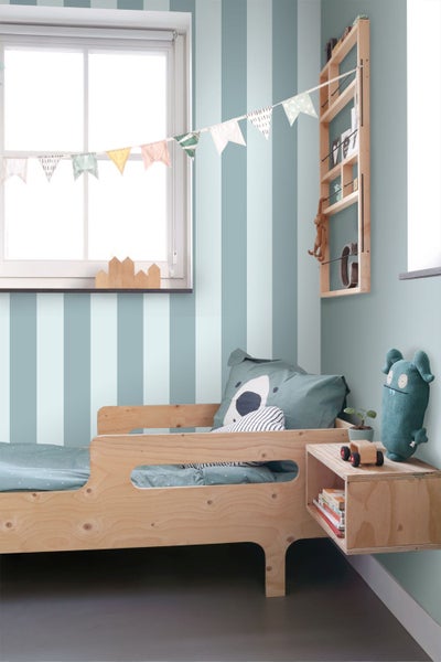 Vlies-Tapete mit breiten vertikalen Streifen in Hellblau und Weiß im Kinderzimmer mit Holzmöbeln und Fenster.