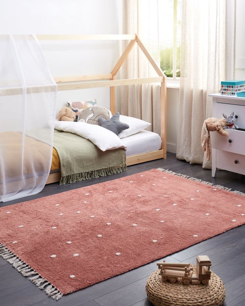 Helles Kinderzimmer mit Hausbett aus Holz, weißer Kommode und einem rosa Teppich mit weißen Punkten und Fransen auf dunklem Boden.