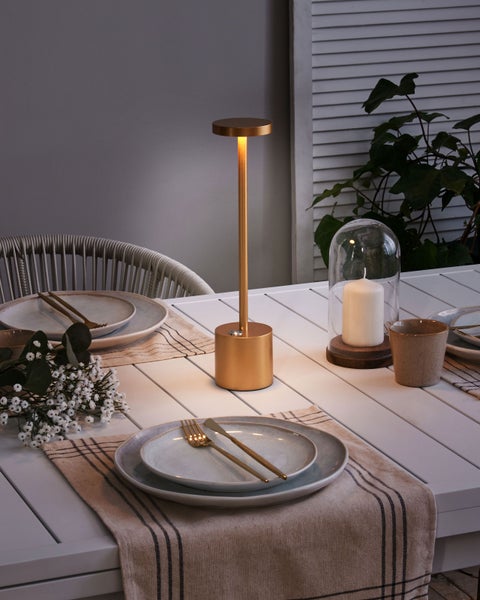 Gedeckter Tisch mit goldfarbener LED Tischleuchte, Geschirr, Kerze unter Glasglocke und Blumendekoration.