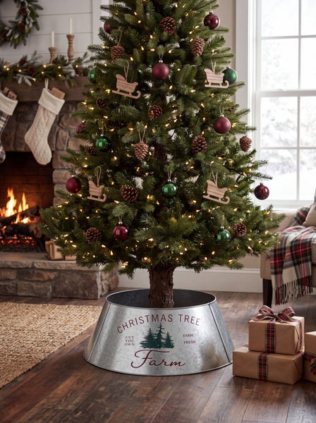 Weihnachtsbaumständer in Metalloptik mit Weihnachtsbaumfarm Aufdruck unter geschmücktem Weihnachtsbaum in einem Wohnzimmer mit Kamin.