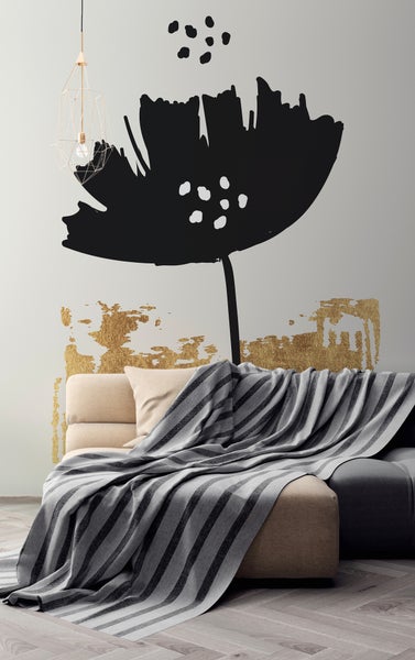 Modernes Wandtattoo mit großer schwarzer Blumensilhouette und goldfarbenen Akzenten an einer Wand hinter einem Sofa mit gestreifter Decke.