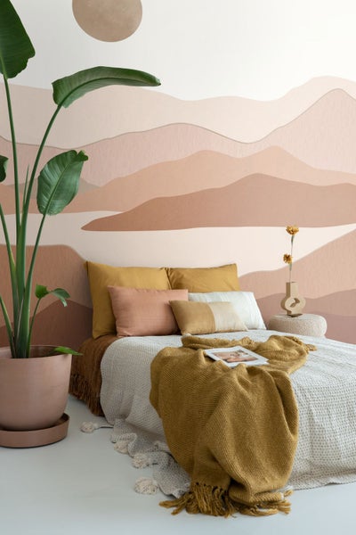 Helles Schlafzimmer mit Bett, Kissen, Decke und Zimmerpflanze im Topf vor einer Wand mit Bergmotiv.