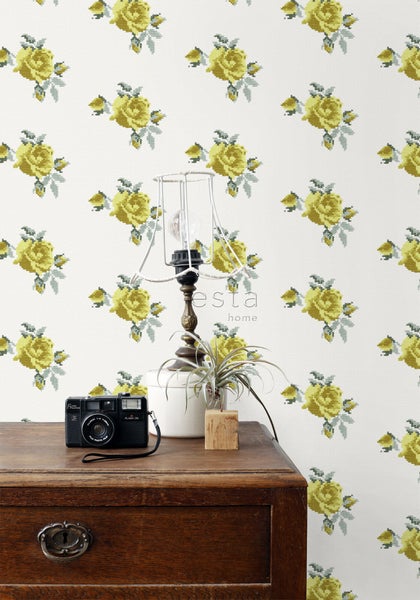 Szene mit Kommode, Lampe, Zimmerpflanze und Kamera vor einer Tapete mit Rosenmuster.