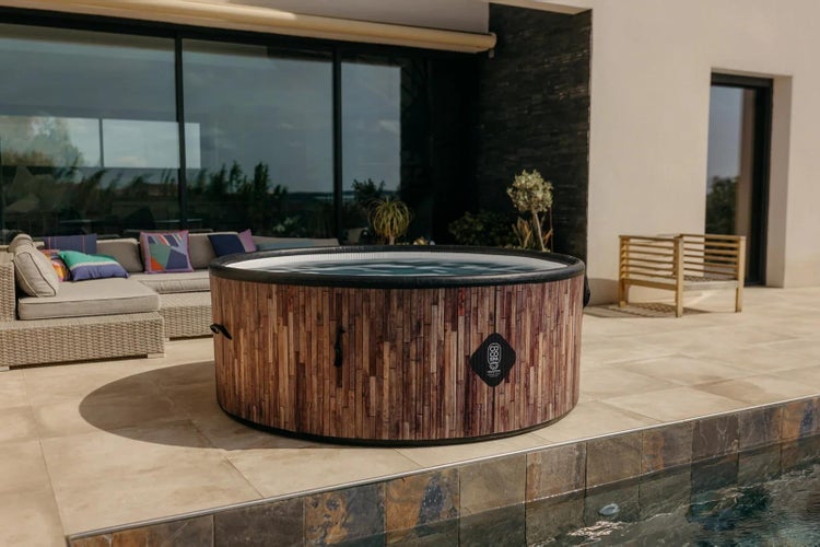 Runder Whirlpool mit Holzoptik und CO SPA Logo auf einer modernen Terrasse neben einem Pool und Gartenmöbeln.