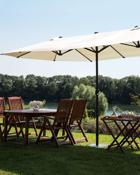 Helle Gartensitzgruppe aus dunklem Holz mit Tisch, vier Klappstühlen, großem weißem Sonnenschirm und Tabletttisch auf einer Wiese an einem See.
