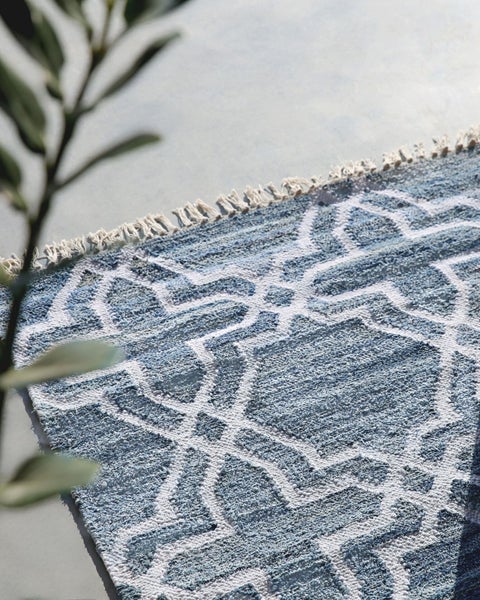 Blau-weiß gemusterter Teppich mit weißen Fransen auf grauem Boden neben einer Pflanze.