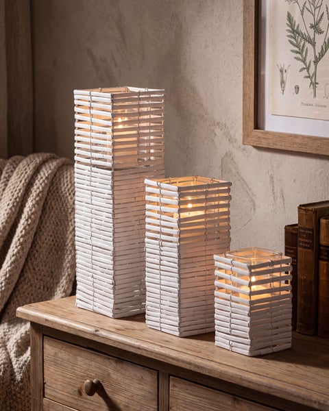 Set aus drei quadratischen Windlichtern aus geflochtenem Material mit Glaseinsatz und brennenden Kerzen auf einer Holzkommode.