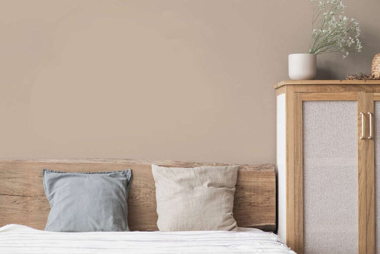 Schlafzimmer mit beige gestrichener Wand, einem Kopfteil aus Naturholz und einem Schrank aus Holz mit Textileinsätzen.