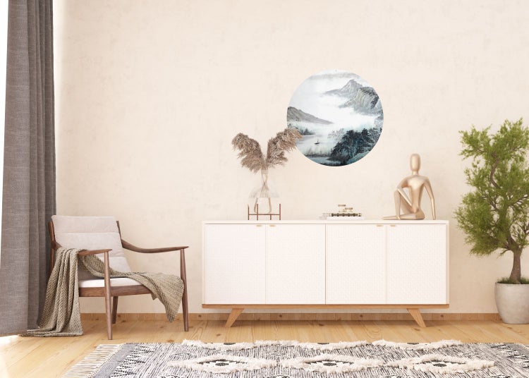 Wohnzimmer mit Sessel, Kommode, Dekorationen und einem runden Wandbild mit Bergmotiven.