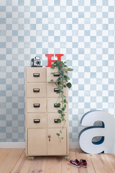 Helle Szene mit karierter Tapete in Hellblau und Weiß, einer Vintage-Kommode aus Holz, Zimmerpflanze und Dekoelementen auf einem Dielenboden.