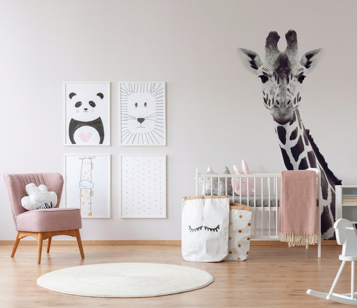 Helles Kinderzimmer mit Giraffen-Fototapete, weißem Gitterbett, rosa Sessel, rundem Teppich und Tierbildern an einer gestreiften Wand.